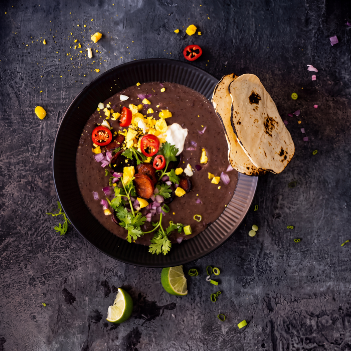 Overhead shot of black bean soup topped with chorizo, egg, chili and cilantro with charred tortillas and lime