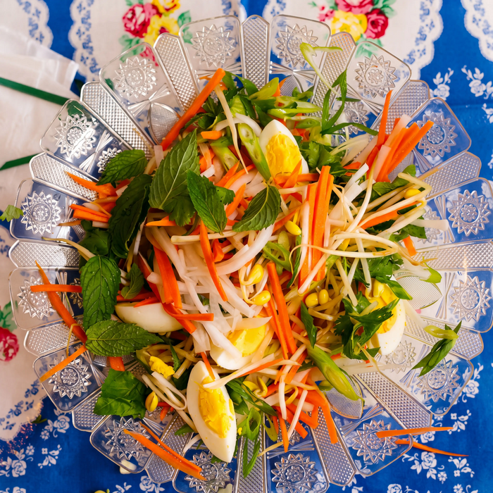 Overhead shot of do chua salad with carrot, daikon, herbs, sprouts and egg in a cut-glass dish on a blue floral tablecloth